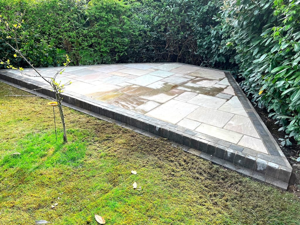 Sandstone patio with a dark block edge set into a lawn beside a young tree