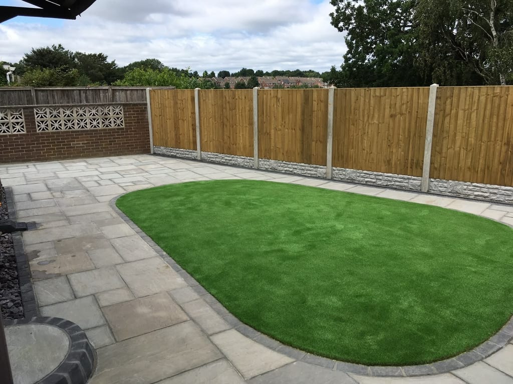 Oval artificial lawn set into grey sandstone paving in front of a decorative concrete screen wall