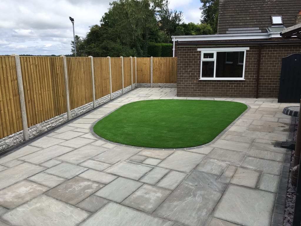 Oval artificial lawn set into grey sandstone paving at the rear of a brown brick bungalow