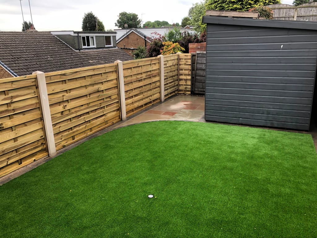 Artificial lawn with a curved sandstone edge beside a grey timber shed and slatted fencing