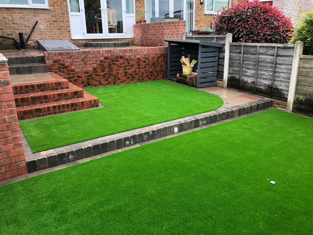Split level artificial lawn with a charcoal block kerb beside a log store and brick steps