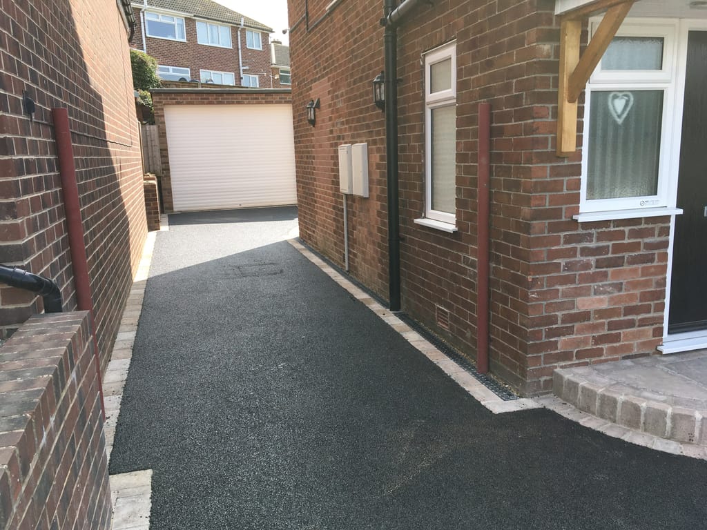 Tarmac driveway running down the side of a red brick house to a white garage door