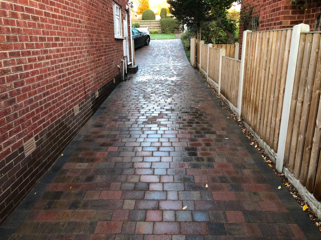 Narrow block paved driveway running between a red brick wall and a close board fence