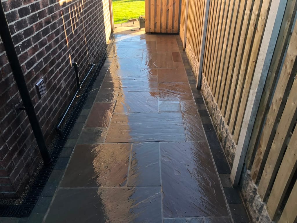 Wet paved passage running between a dark brick house wall and a vertical timber fence