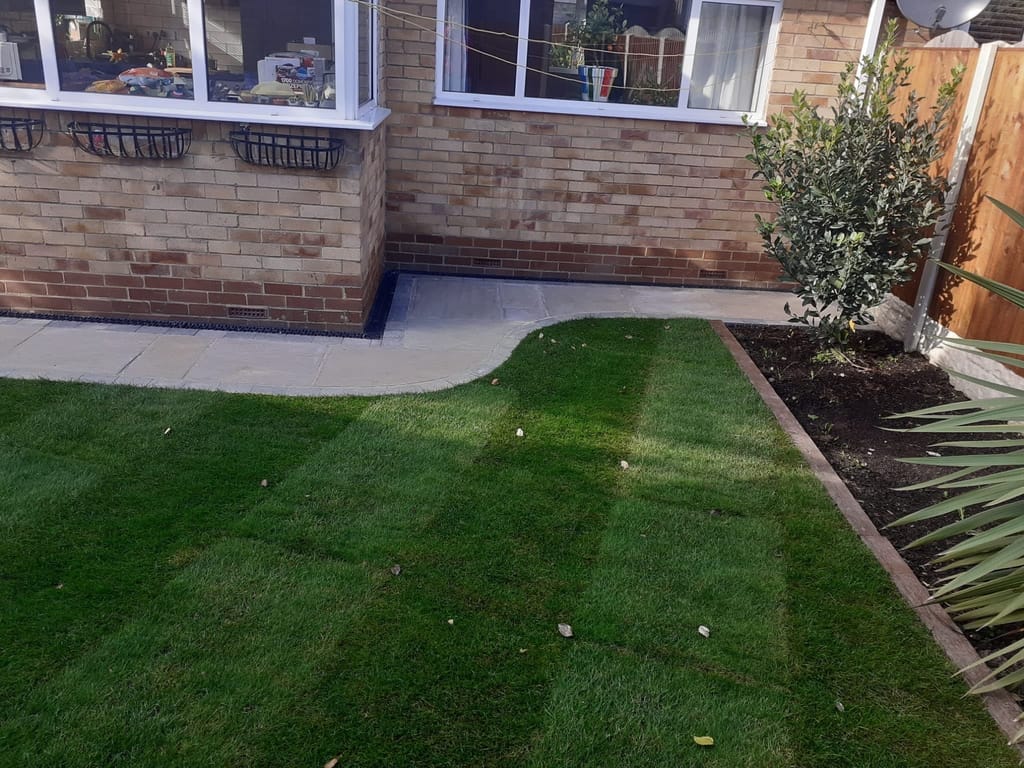 Buff slab path with a curved edge meeting freshly laid turf behind a brick house