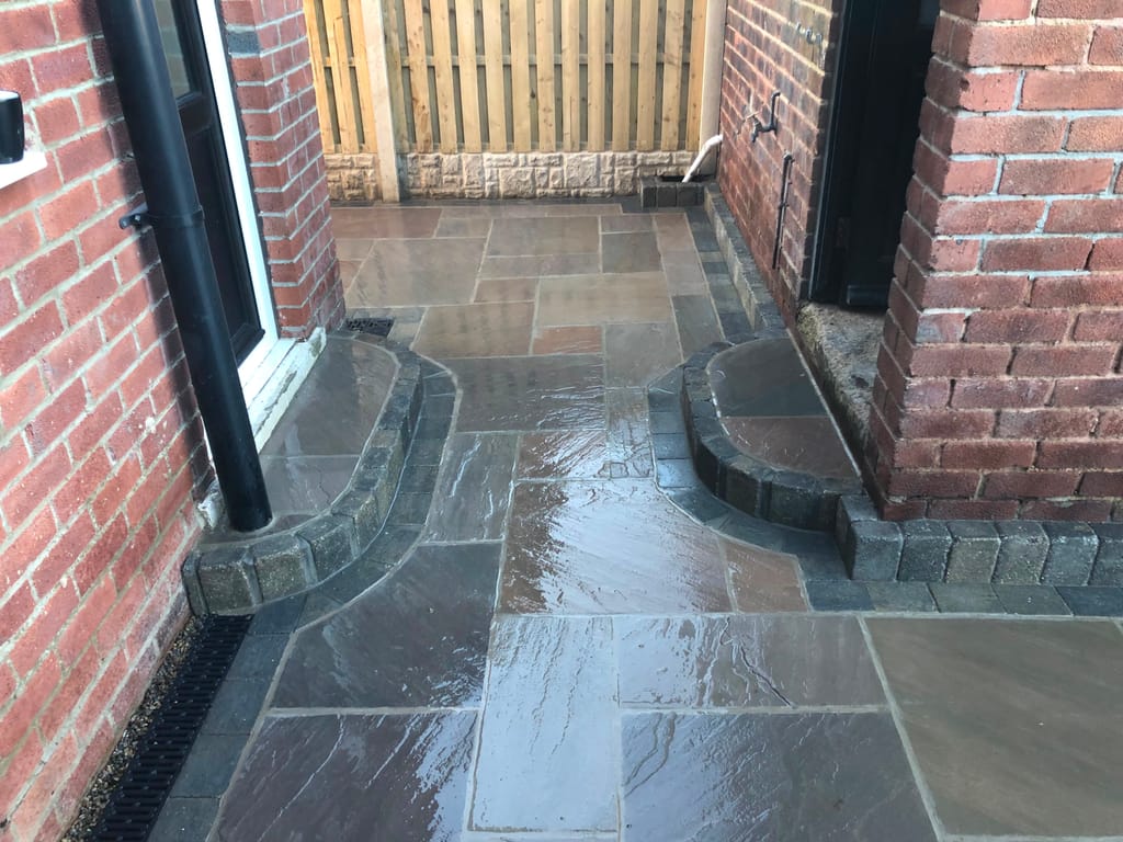 Wet sandstone paving between red brick walls with two curved sett edged doorsteps