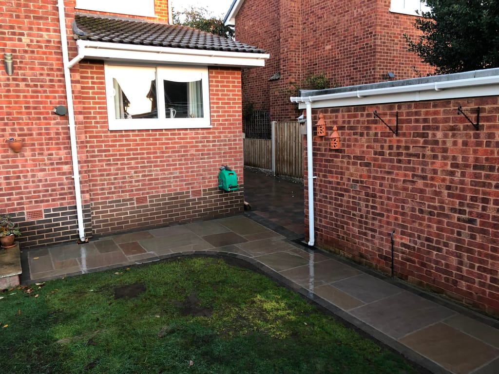 Sandstone paving turning the corner of a red brick house beside a lawn and a garage wall