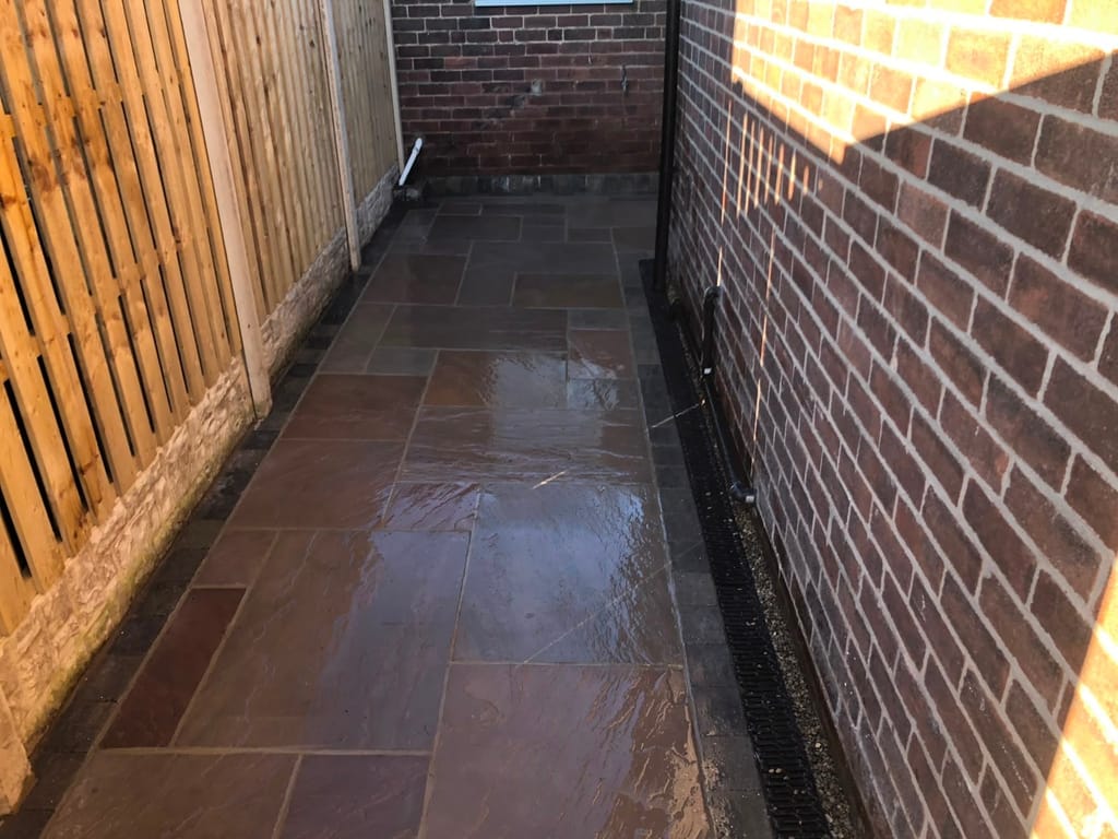 Wet sandstone side path with a channel drain between a red brick wall and a timber fence