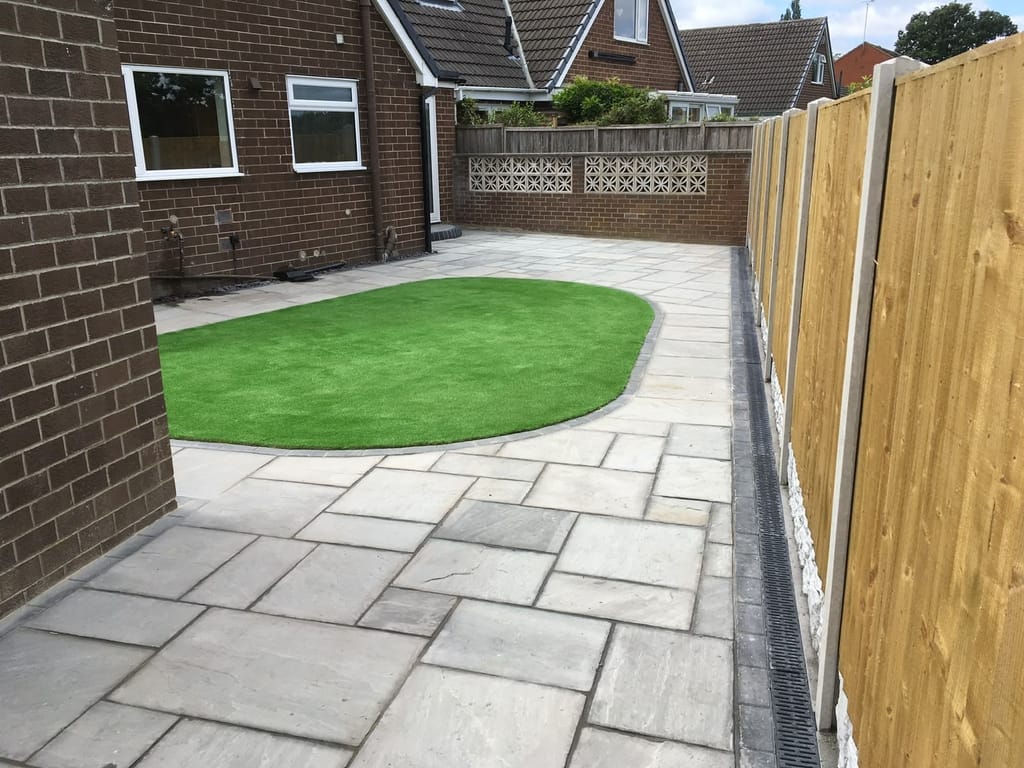 Grey sandstone patio wrapping round an oval artificial lawn beside a dark brick bungalow