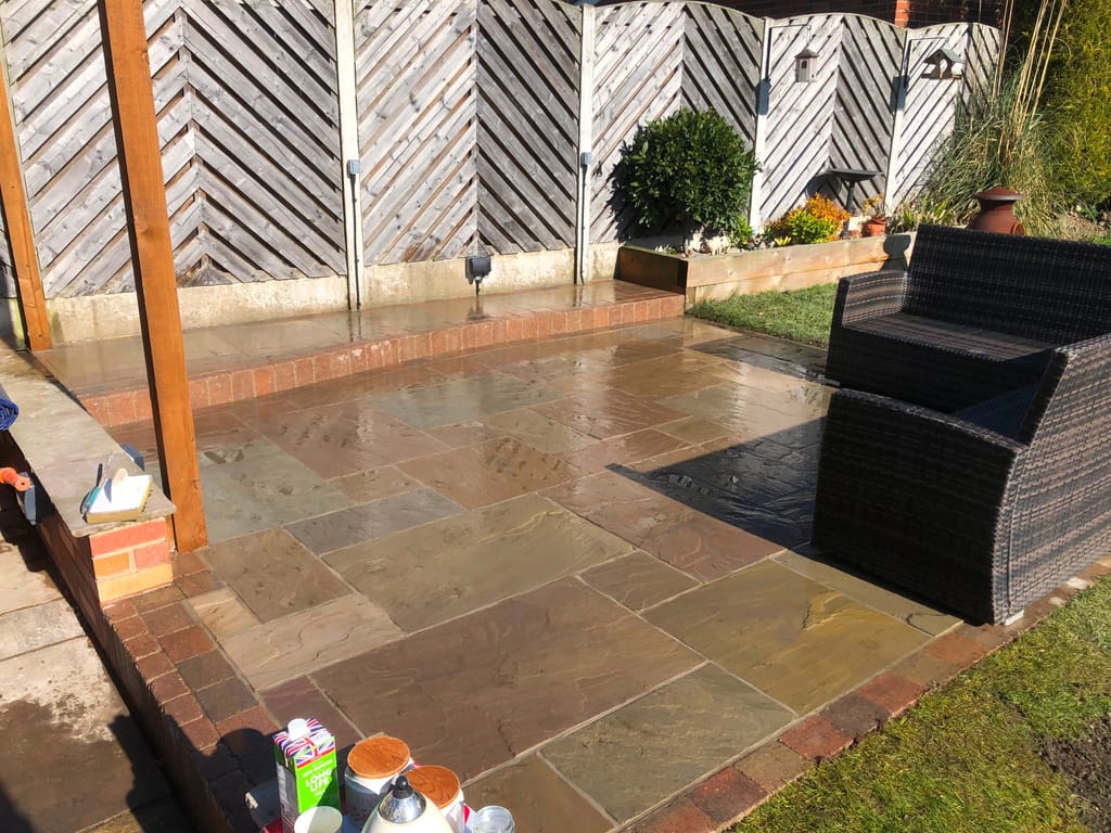 Sandstone patio with brick edging under a timber pergola beside chevron fence panels