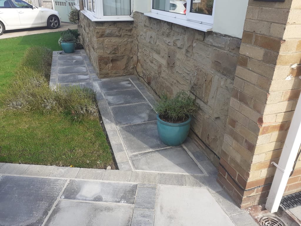 Dark slab paving running round the stone front of a bungalow past a lavender bed