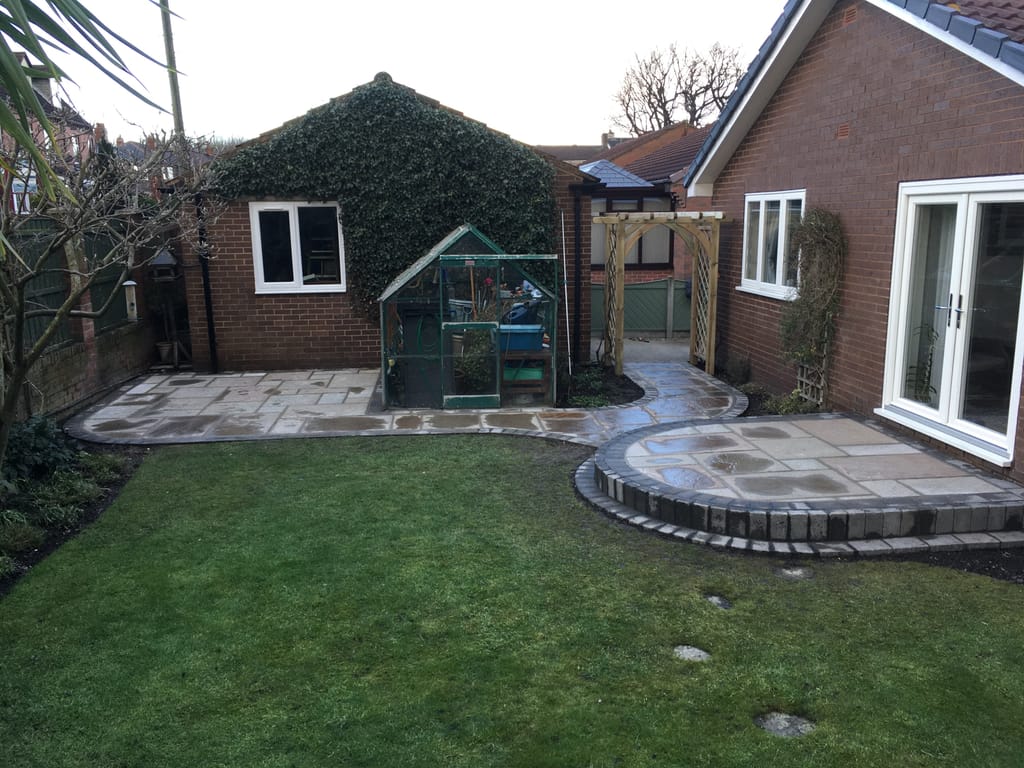 Sandstone patio with a circular sett edged bay at french doors beside a greenhouse and a timber arch
