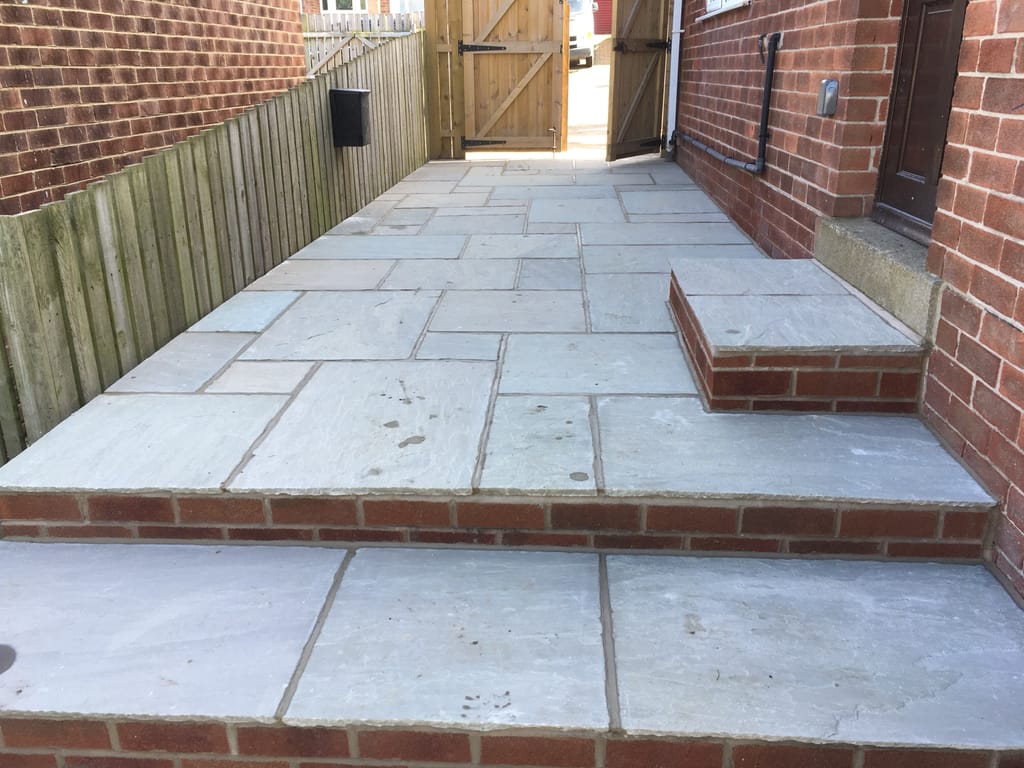 Grey sandstone paving raised on a brick course beside a timber fence and a side gate