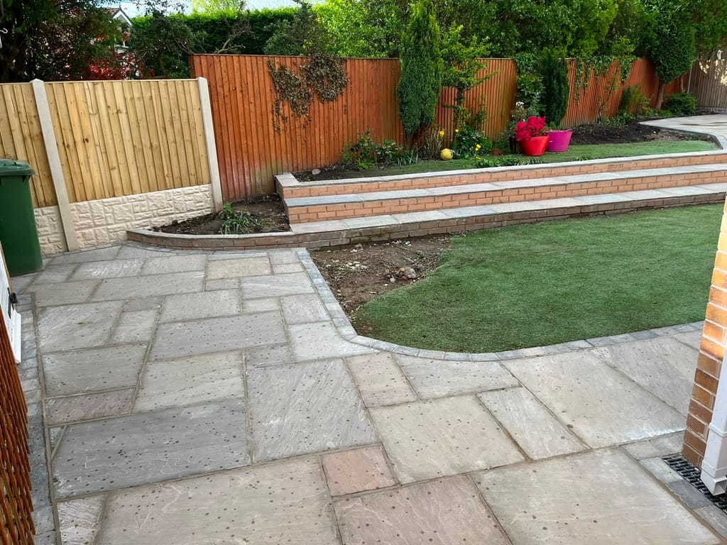 Sandstone patio below a red brick retaining wall holding a raised artificial lawn