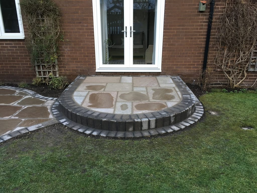 Curved sandstone landing with a charcoal sett riser outside the french doors of a red brick bungalow